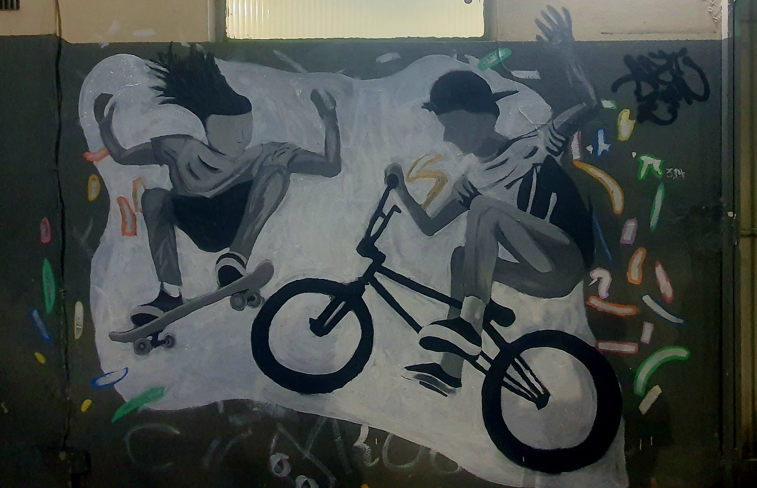 Mural em estilo grafite retratando duas figuras em tons de cinza praticando esportes radicais. À esquerda, uma pessoa salta com um skate, com o cabelo estilizado em movimento. À direita, outra pessoa realiza uma manobra em uma bicicleta BMX. Ao redor das figuras, há formas coloridas abstratas e o fundo é predominantemente escuro com detalhes em branco.