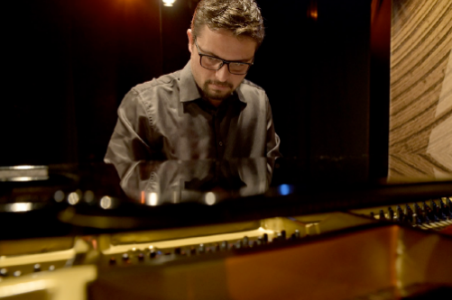 Homem com barba e óculos, usando uma camisa social escura, toca piano de cauda em ambiente com iluminação suave e decoração de madeira. Ele está concentrado, olhando para as teclas, e seu reflexo é visível na superfície brilhante do piano.
