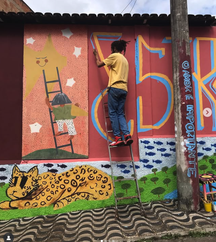 Pessoa em pé numa escada pintando um mural colorido em uma parede externa. A arte inclui uma grande estrela sorridente com uma criança subindo uma escada em sua direção, várias estrelas pequenas, e um grande felino amarelo com manchas pretas deitado sobre grama verde, próximo a uma árvore. À direita, o artista pinta letras grandes em amarelo com contorno azul. Um poste de concreto na frente da parede tem a frase "O AMOR É IMPORTANTE" escrita em azul. No chão há um balde com pincéis e latas de tinta.