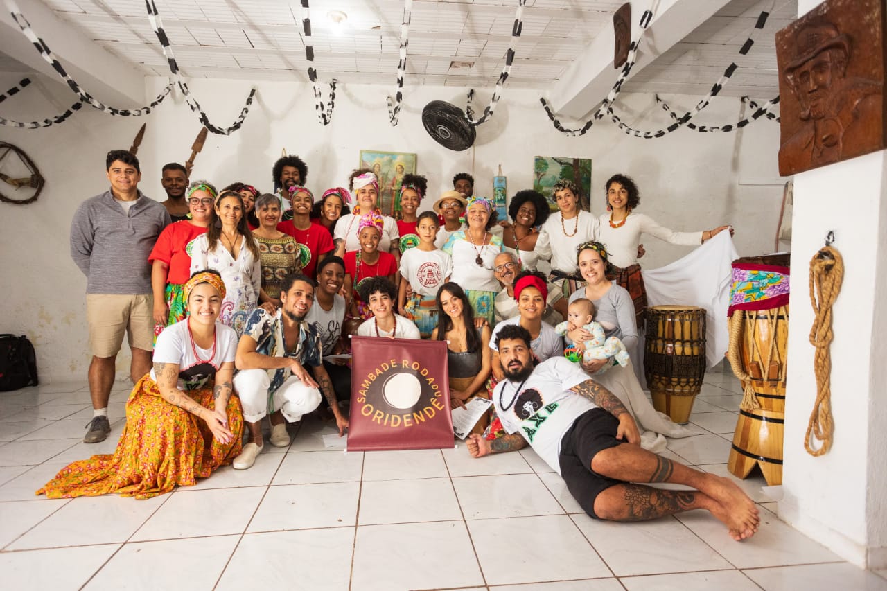 Um grupo diverso de aproximadamente 30 pessoas posa sorridente para uma foto em um ambiente interno decorado com correntes de papel penduradas no teto. Muitas pessoas usam roupas coloridas, saias estampadas e turbantes, refletindo elementos da cultura afro-brasileira. Ao centro, duas pessoas seguram um estandarte com os dizeres "Samba de Roda de Oridendê". Há instrumentos de percussão, como atabaques, visíveis ao lado direito da imagem. A atmosfera é alegre e comunitária, sugerindo um evento cultural.