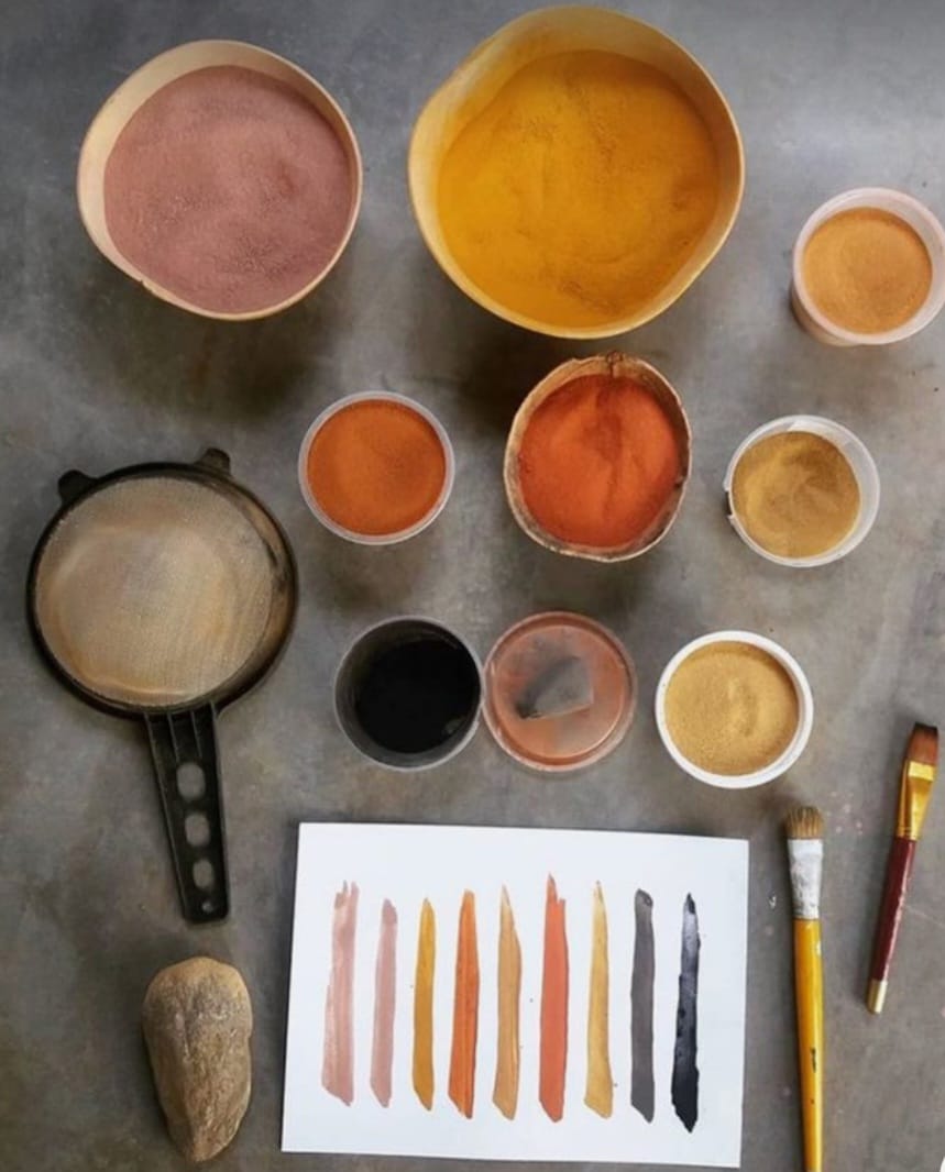 Vista superior de recipientes com pigmentos naturais em tons terrosos, como amarelo, ocre, vermelho, laranja, marrom e preto, dispostos sobre uma superfície de concreto. Abaixo, há um papel com amostras de tinta feitas com esses pigmentos, pincéis, uma peneira, uma pedra e um pequeno recipiente com líquido escuro, possivelmente aglutinante ou tinta. A imagem sugere um processo artesanal de produção de tintas naturais.
