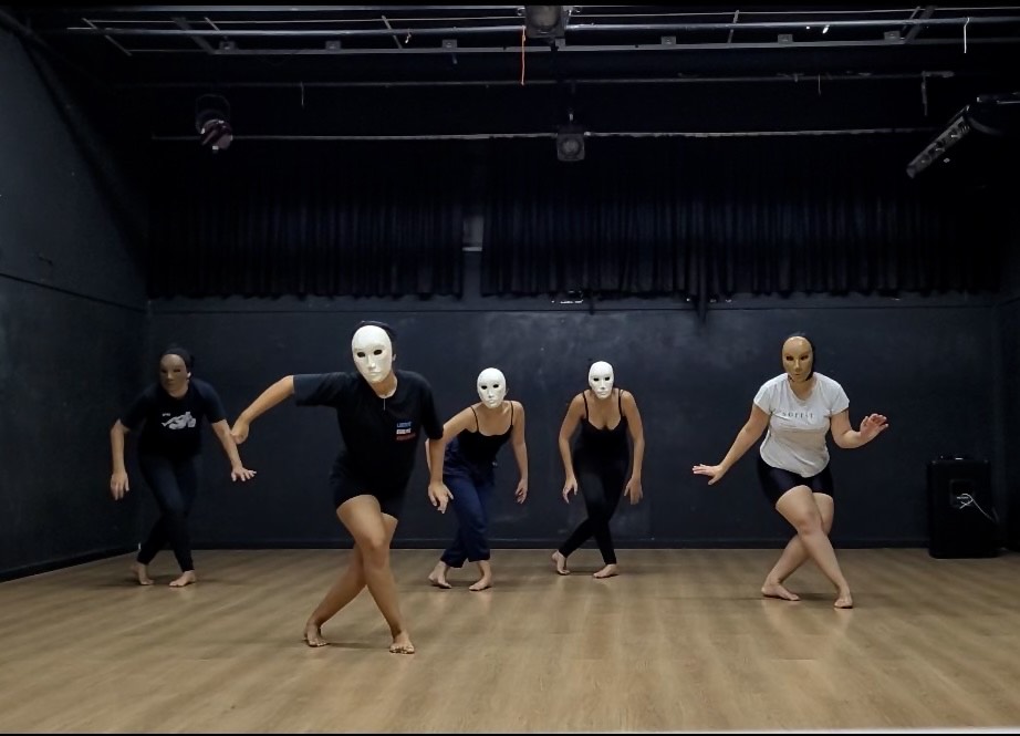 Cinco pessoas com máscaras brancas se apresentam em um estúdio de dança de piso de madeira e paredes escuras. Elas estão descalças e vestem roupas pretas ou escuras, exceto uma pessoa à direita que usa camiseta branca. Estão em posições coreografadas, com posturas corporais expressivas e sincronizadas, transmitindo intensidade e concentração. O ambiente é fechado, com iluminação controlada e equipamentos de som visíveis ao fundo.