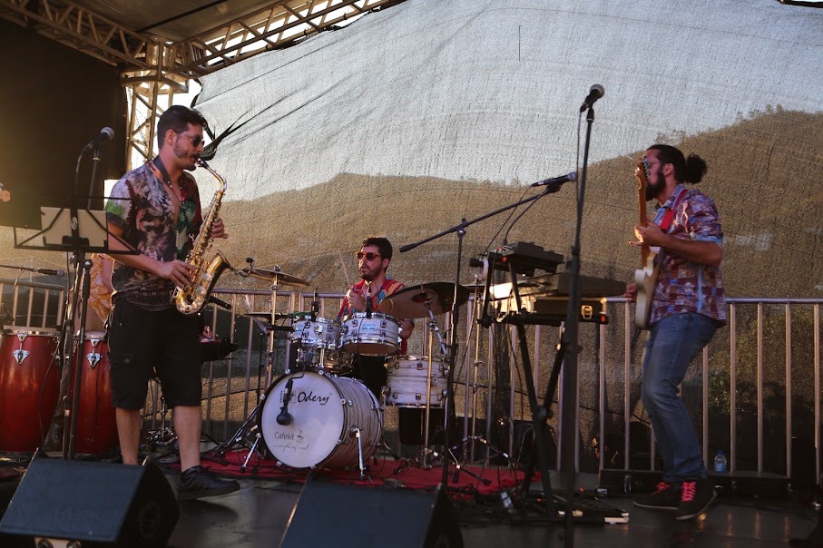 Banda se apresentando ao vivo em um palco ao ar livre durante o dia. À esquerda, um músico toca saxofone alto; no centro, o baterista está tocando uma bateria branca com a marca "Odery"; à direita, outro músico toca contrabaixo elétrico. Todos usam roupas casuais coloridas e óculos escuros. Ao fundo, há uma lona preta cobrindo parcialmente o palco, com uma paisagem de morros visível através dela. Instrumentos adicionais como teclados e congas também estão no palco.
