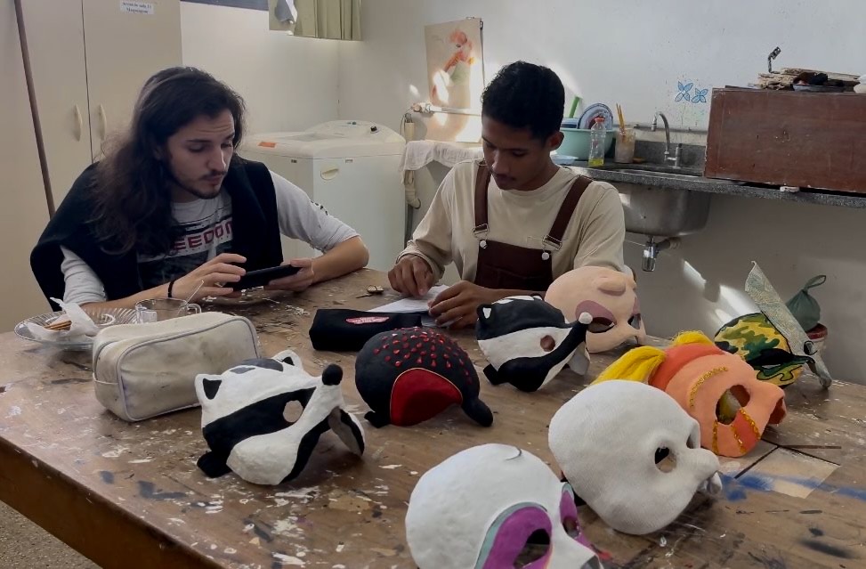 Dois jovens estão sentados em uma mesa de ateliê, concentrados em atividades manuais e usando materiais artísticos. Um deles olha para um celular enquanto o outro escreve em um papel. Sobre a mesa há diversas máscaras artesanais coloridas, com formas e estilos variados, além de estojos e pincéis. Ao fundo, é possível ver uma pia com utensílios de pintura e armários. A cena se passa em um ambiente de criação artística.