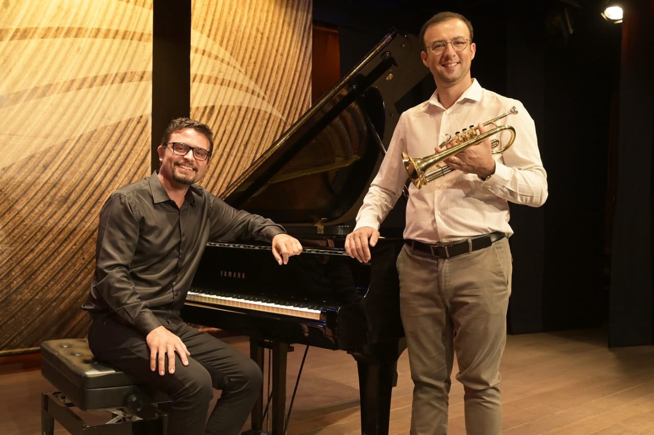 Dois músicos posam sorridentes em um palco com fundo de madeira texturizada; o da esquerda está sentado ao piano de cauda Yamaha, vestindo camisa preta e óculos, enquanto o da direita está de pé segurando um trompete dourado, usando camisa clara e calça bege.