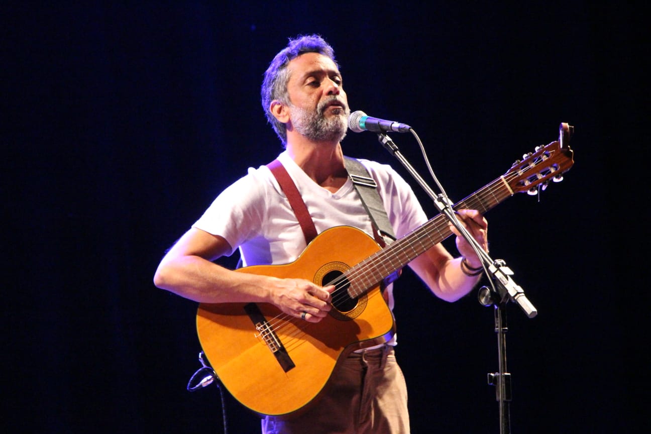 Homem de barba grisalha toca violão e canta em um microfone durante apresentação em palco escuro, iluminado por luz azul. Ele veste camiseta branca, suspensórios bordô e calça bege, com expressão concentrada enquanto canta e toca. Dois microfones estão posicionados à sua frente.