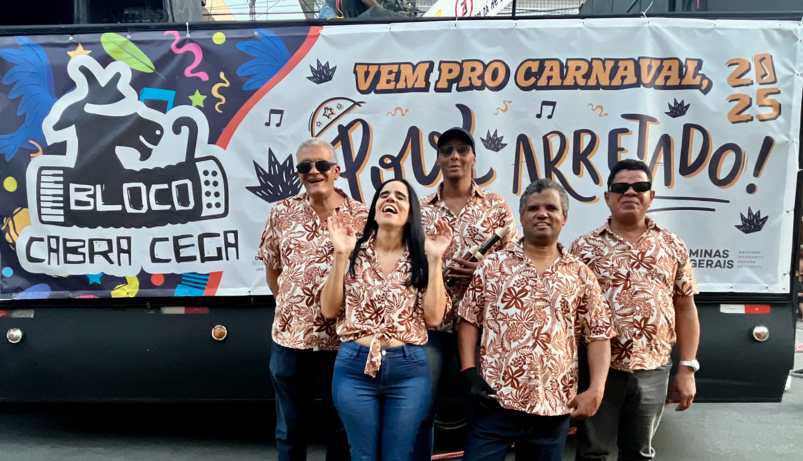 Cinco pessoas posam sorrindo em frente a um banner colorido do bloco de carnaval “Bloco Cabra Cega”. Todos vestem camisas floridas em tons de marrom e bege. O banner ao fundo tem elementos festivos como notas musicais, confetes e folhas, e os dizeres “Vem pro Carnaval 2025 – Puxado Arretado!”. As pessoas estão felizes e em clima de celebração, indicando um evento carnavalesco em Minas Gerais.