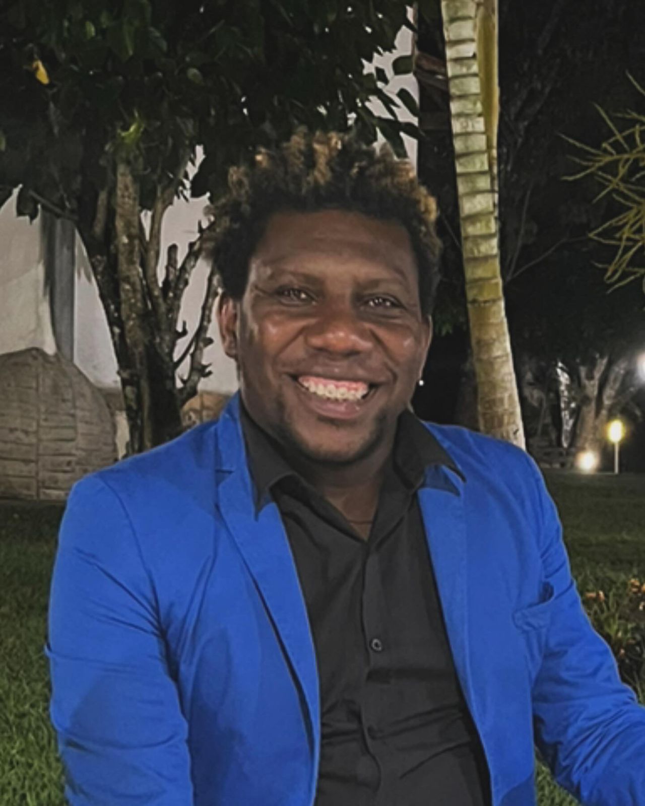 Homem sorridente usando blazer azul e camisa preta, sentado ao ar livre à noite, com árvores e iluminação suave ao fundo.