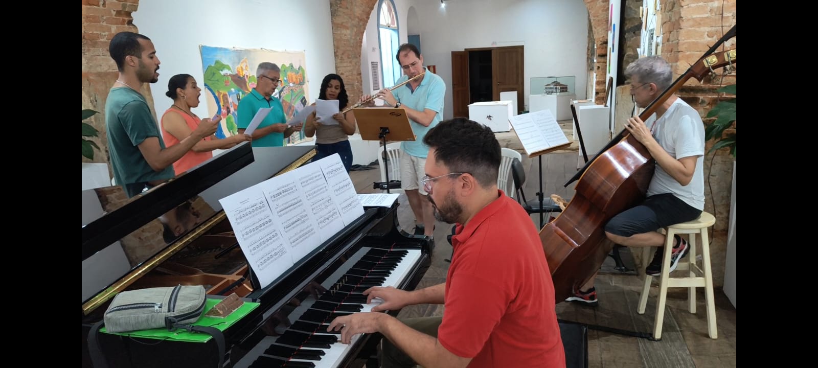 Ensaio musical em ambiente interno com paredes de tijolos aparentes e arcos de pedra. À esquerda, cinco pessoas em pé cantam em coro, segurando partituras. À direita, um músico toca flauta transversal e outro toca contrabaixo acústico sentado em um banquinho. Em primeiro plano, um pianista de camisa vermelha toca um piano de cauda com partituras abertas à sua frente. O espaço tem iluminação natural e clima descontraído, com obras de arte ao fundo e mobiliário simples.
