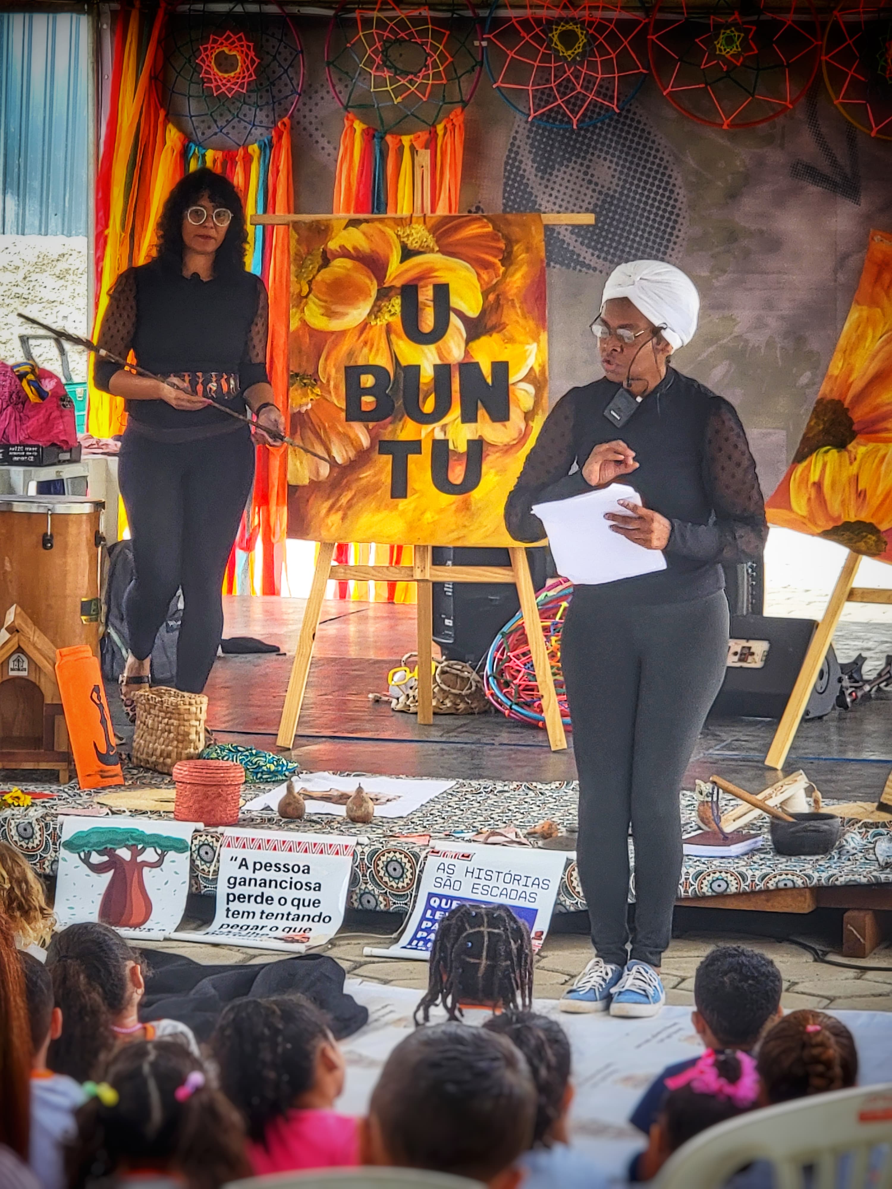 Na foto, vemos uma mulher negra, com turbante branco, de pé, falando no microfone. Atrás dela, há um cartaz com o texto "UBUNTU" e outra mulher negra à esquerda. Há várias crianças sentadas à frente, assistindo, viradas para ambas as mulheres.