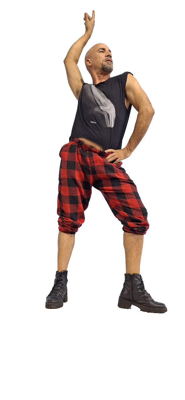 Fotografia em fundo branco de um homem careca com cavanhaque em pose artística. Ele está com as pernas afastadas, uma mão na cintura e o outro braço erguido de forma elegante, olhando para o alto. Ele veste uma camiseta preta sem mangas com uma estampa cinza, calças largas com estampa xadrez vermelha e preta na altura das canelas e botas pretas.