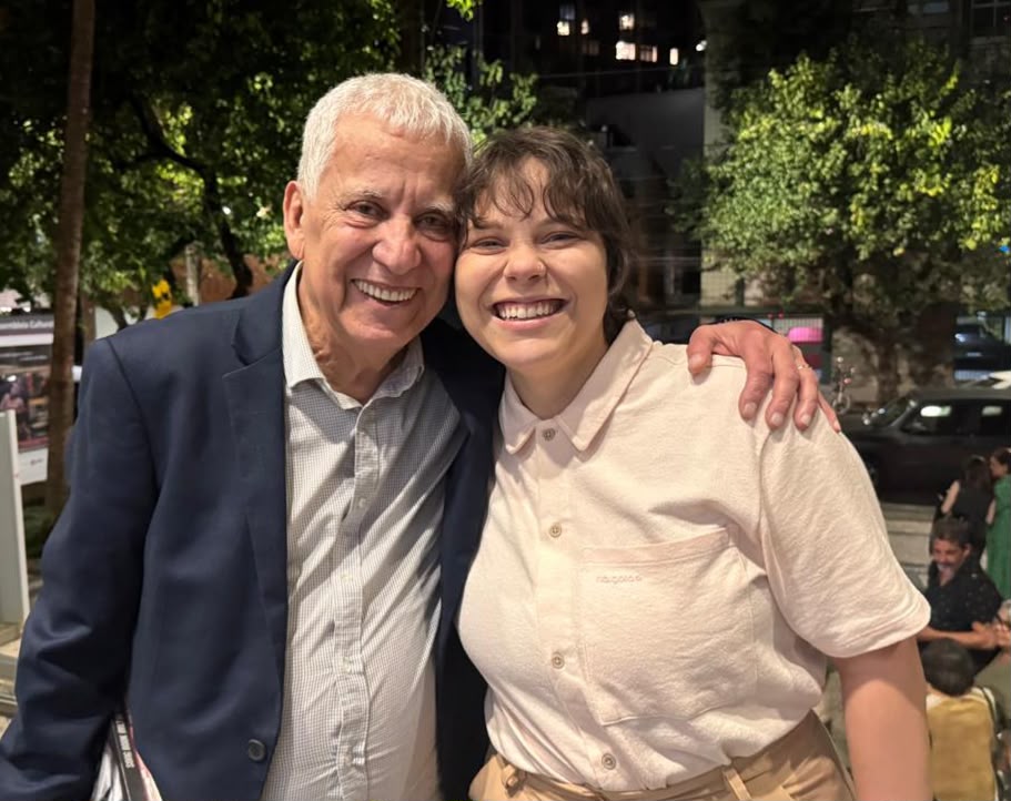 Fotografia de Márcio Borges e Helena Borges Fotografia noturna ao ar livre de duas pessoas abraçadas e sorrindo para a câmera. À esquerda, um homem idoso de cabelos brancos, vestindo paletó azul e camisa clara, está com o braço no ombro da pessoa ao lado. À direita, uma jovem de cabelos curtos escuros veste uma camisa clara de botões. Ao fundo, um pouco desfocados, há árvores, rua, carros e pessoas em uma calçada.