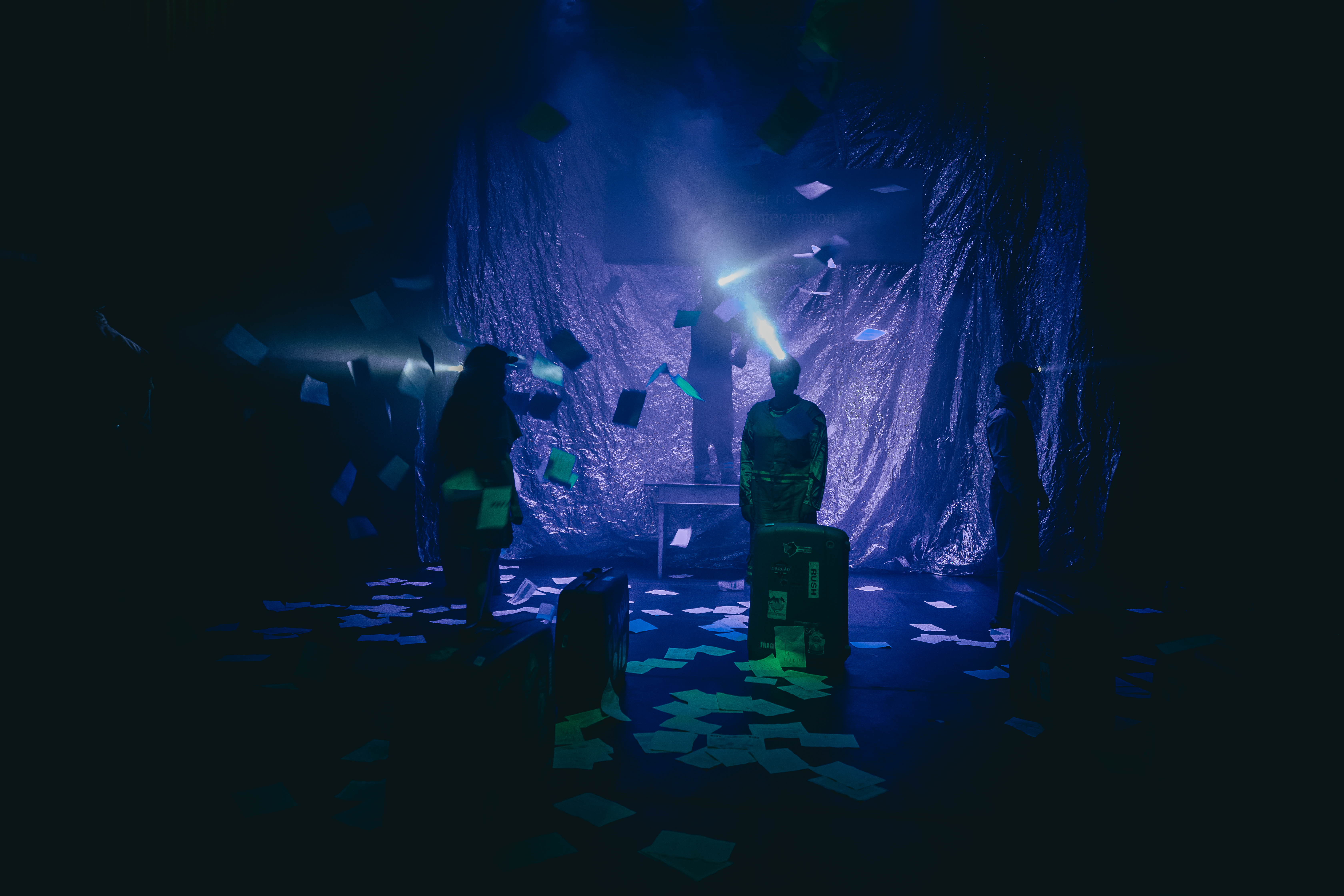 Cena de performance com luzes e papéis flutuando Cena de performance teatral em um palco escuro com luz azul. O fundo é uma cortina reflexiva. Inúmeros papéis estão espalhados pelo chão e suspensos no ar, com brilhos esverdeados. Três performers são vistos como silhuetas. Duas delas têm lanternas de cabeça projetando feixes de luz. A figura central está em pé sobre malas e uma mesa. Malas de viagem estão pelo palco. Atmosfera dramática e misteriosa.