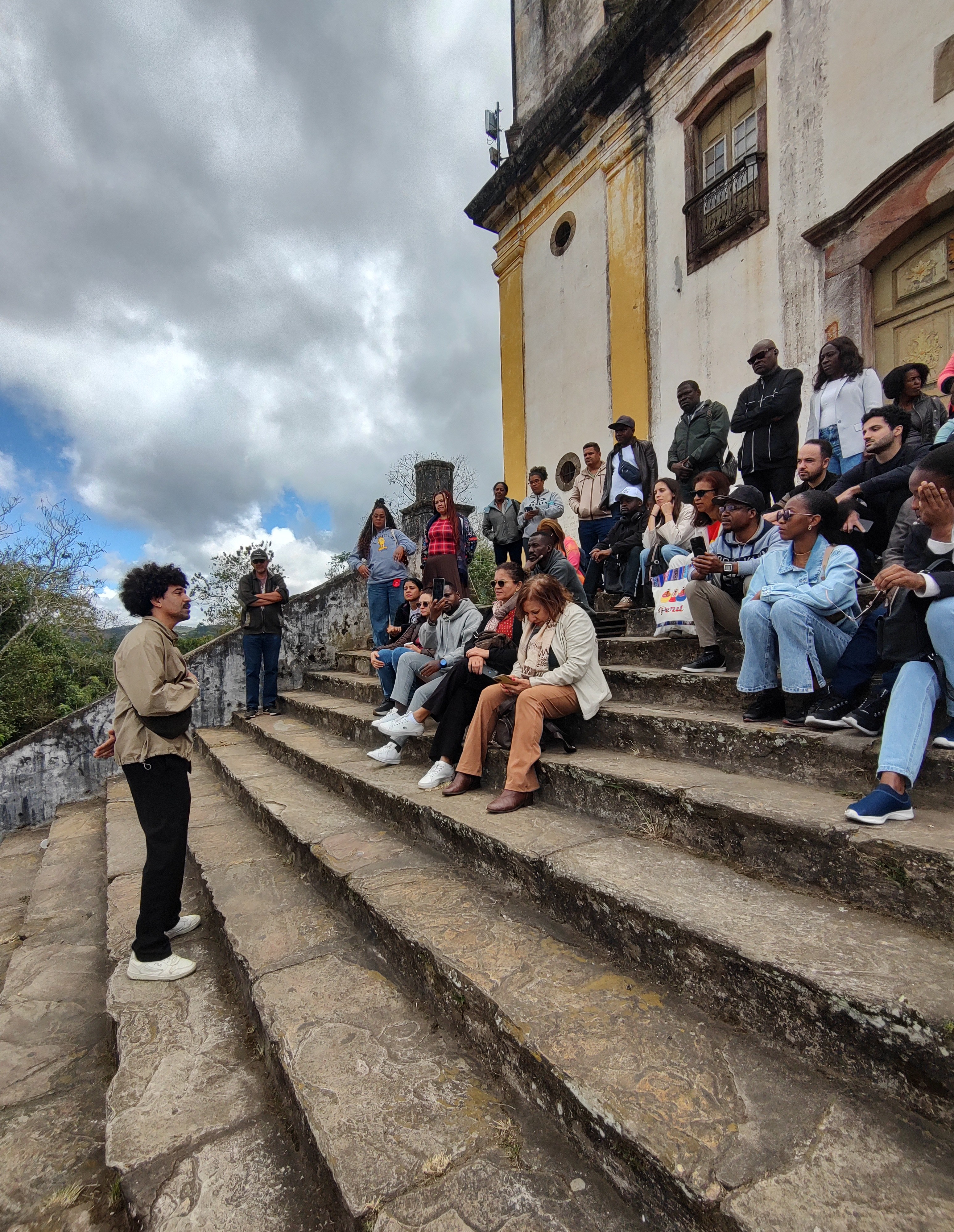Apresentação Palma Preta: en(canto) ancestral Fotografia ao ar livre. À esquerda, um homem negro com cabelo crespo, vestindo jaqueta bege e calça preta, está em pé nos degraus de pedra, falando para um grupo. À direita, dezenas de pessoas estão sentadas e em pé na ampla escadaria de pedra de uma igreja colonial antiga, de paredes brancas e amarelas, prestando atenção nele. O céu ao fundo está nublado com aberturas azuis.