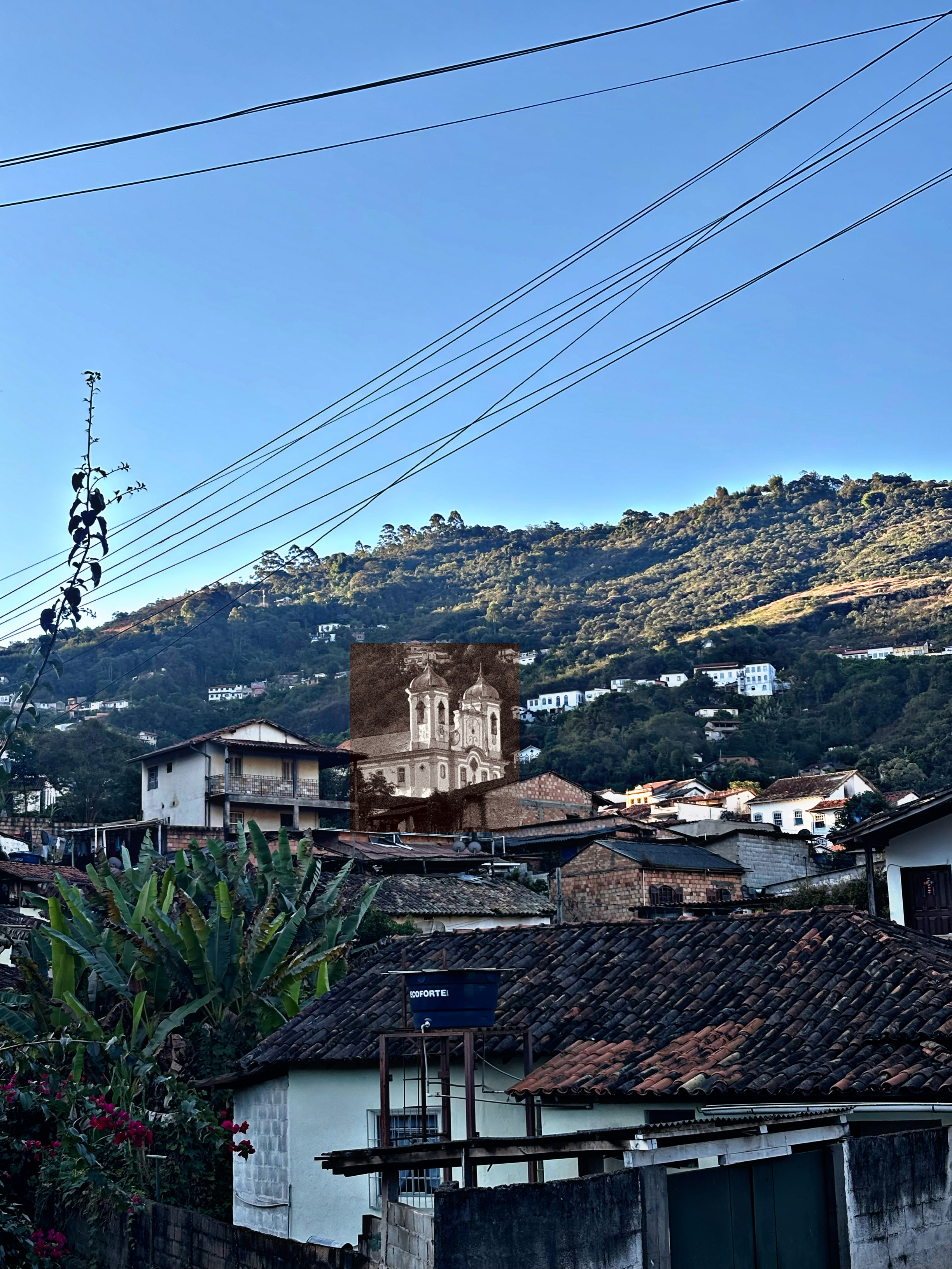 Fotografia de uma paisagem urbana. Em primeiro plano, telhados de barro de casas simples, uma caixa d'água, folhagens e fios de eletricidade cruzando um céu azul claro. Ao fundo, uma montanha verde salpicada de casas. No centro da cena, destacando-se das construções, há uma inserção retangular em tom sépia mostrando uma igreja colonial de duas torres, contrastando com o restante colorido da foto.