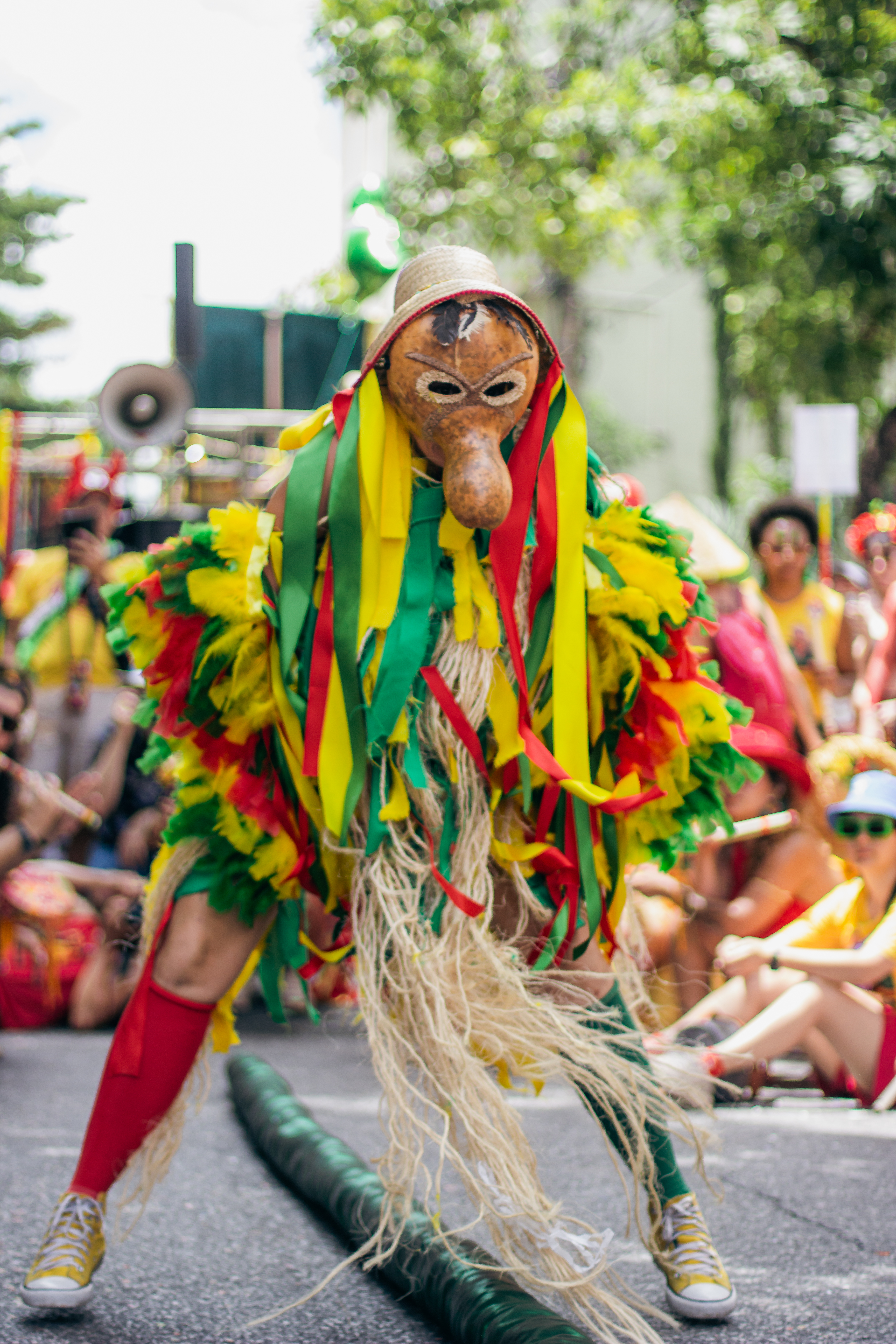 Caretos Fotografia colorida de um artista com traje performático agachado no centro de uma rua de asfalto. A pessoa usa uma máscara de cabaça rústica com nariz longo e um pequeno chapéu de palha. O traje é vibrante, coberto por franjas de tecido amarelas, verdes e vermelhas e fibras naturais longas. Usa tênis e meias altas coloridas. Ao fundo, uma multidão desfocada assiste sob árvores. No chão, um longo tubo verde.