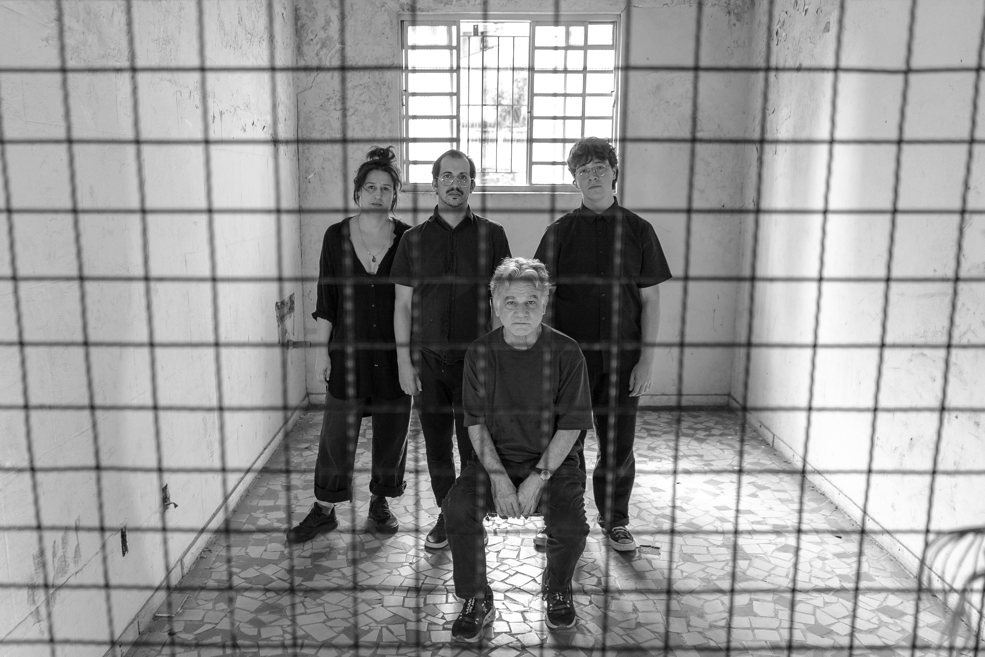 Fotografia da equipe em preto e branco Fotografia em preto e branco de quatro pessoas em uma sala vazia, vista através de uma grade de arame em primeiro plano. Um homem mais velho está sentado no centro. Atrás dele, três pessoas em pé, sendo uma mulher à esquerda e dois homens à direita. Todos vestem roupas escuras e têm expressões sérias. O chão é de piso de caquinhos e, ao fundo, há uma janela com grades.